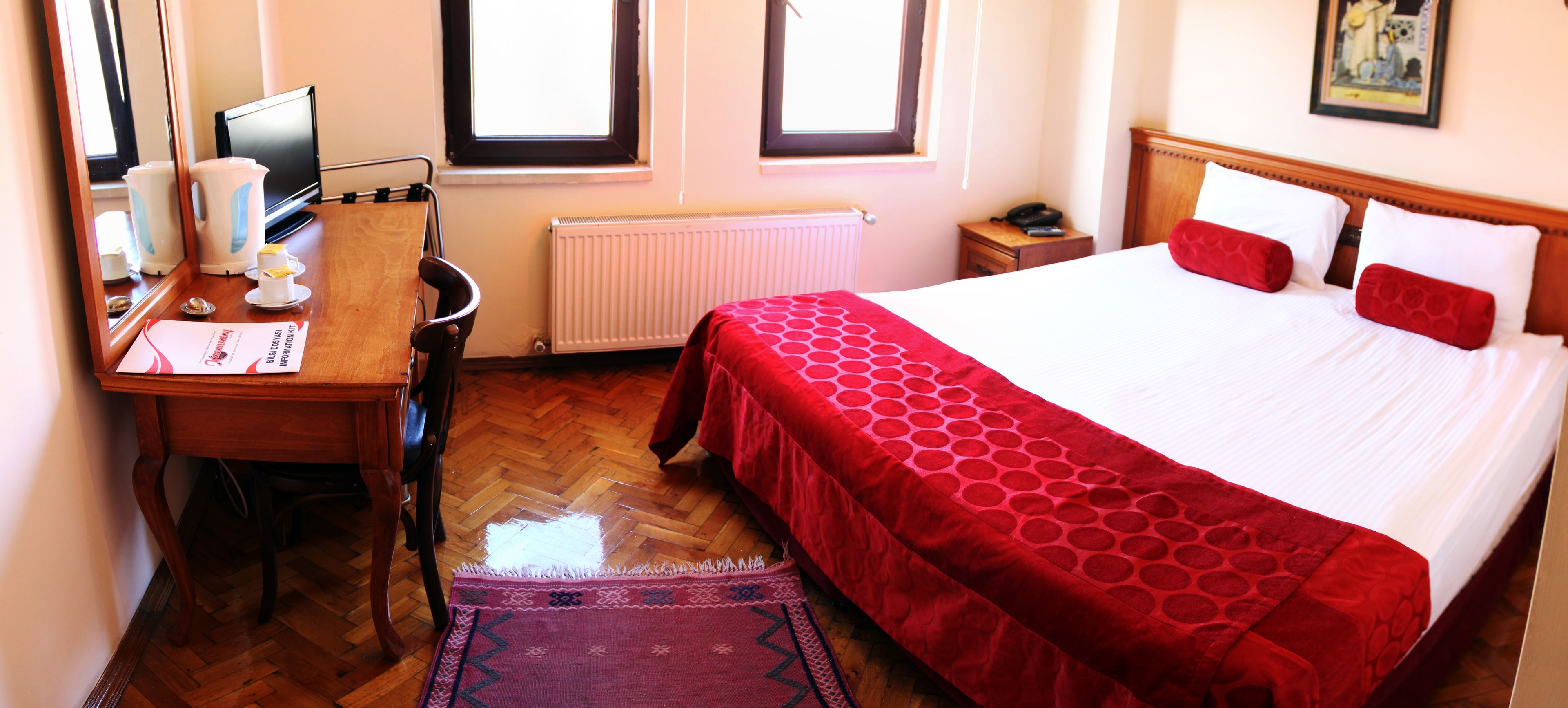 A hotel room with a bed, a desk with a chair, a radiator, and a window.