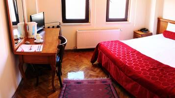 A hotel room with a bed, a desk with a chair, a radiator, and a window.