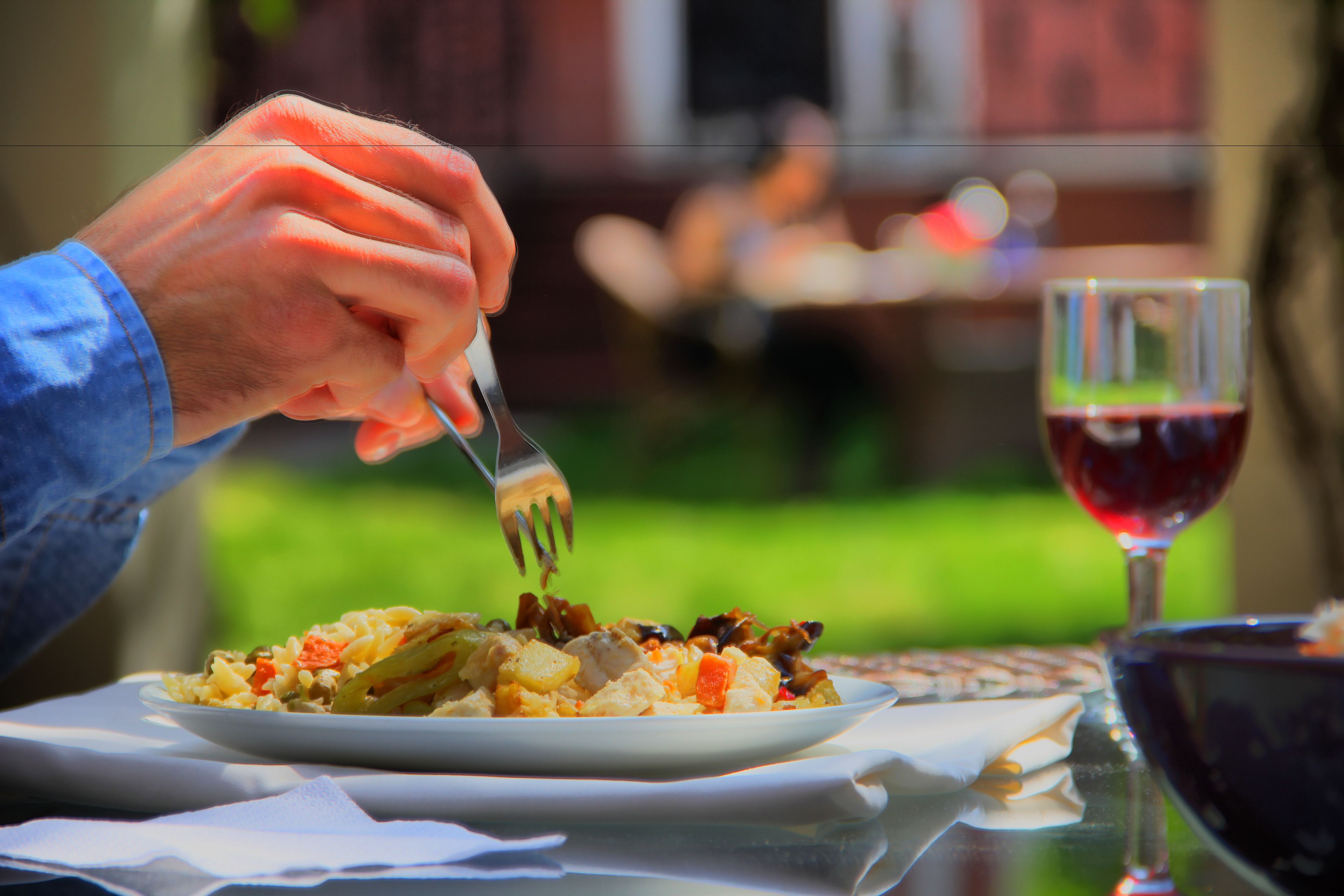 A person dining outdoors with a plate of food and a glass of red wine on a table.