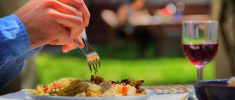 A person dining outdoors with a plate of food and a glass of red wine on a table.