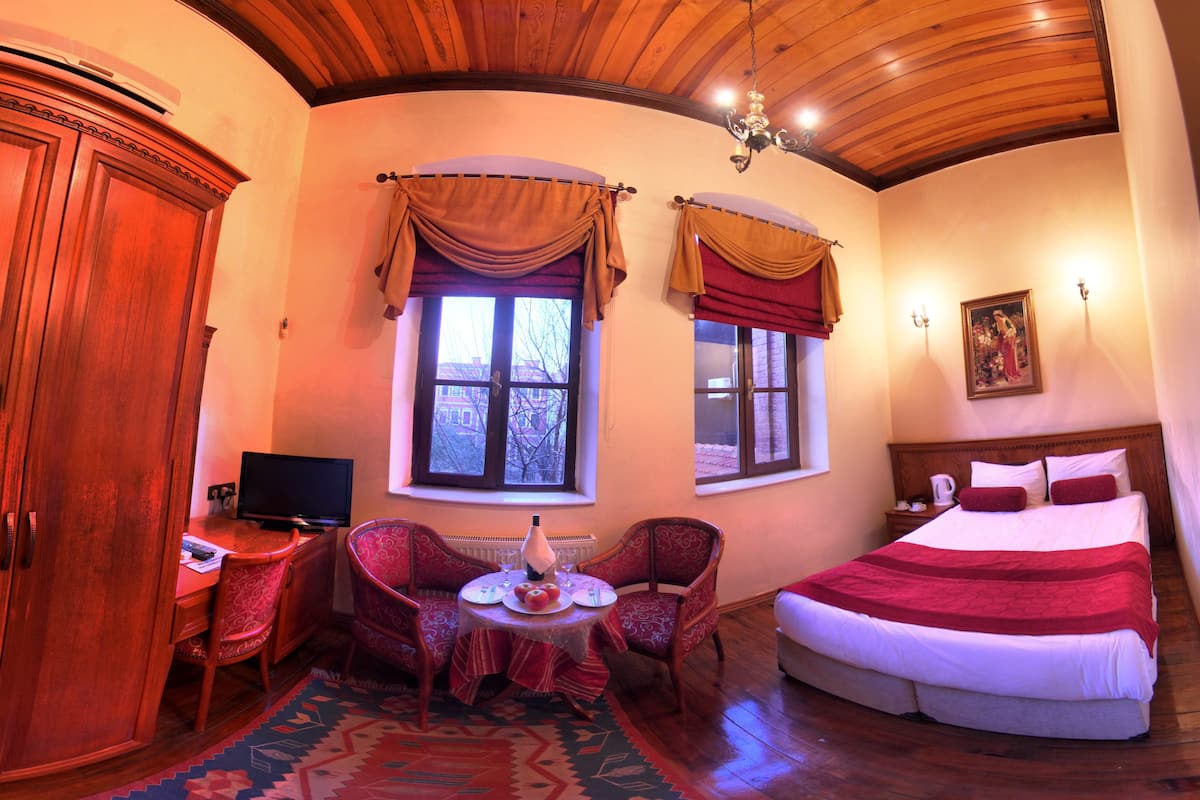 A hotel room with a wooden ceiling, a bed with red bedding, two windows with curtains, a television, a small table with a lamp, and a patterned rug.