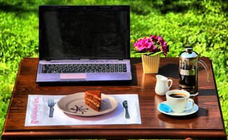 Una laptop, una taza de café, una porción de pastel, una cuchara, una jarra y una maceta con una flor sobre una mesa de madera al aire libre.