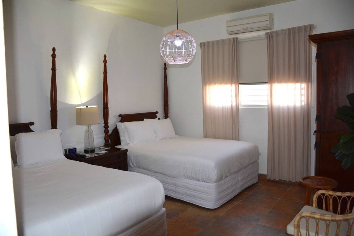 Quarto de hotel com duas camas de solteiro, uma mesa de cabeceira com abajur, uma janela grande com cortinas e um guarda-roupa de madeira.