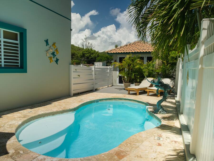 En swimmingpool med en hvid fliseterrasse, hvide hegn og en bygning med blå vinduesramme.