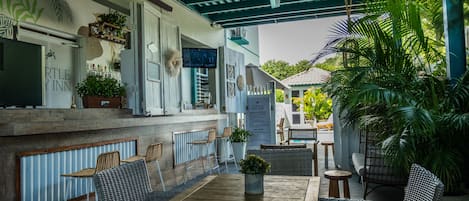 A cozy outdoor seating area with wicker furniture and a wooden table, surrounded by potted plants and a blue ceiling.