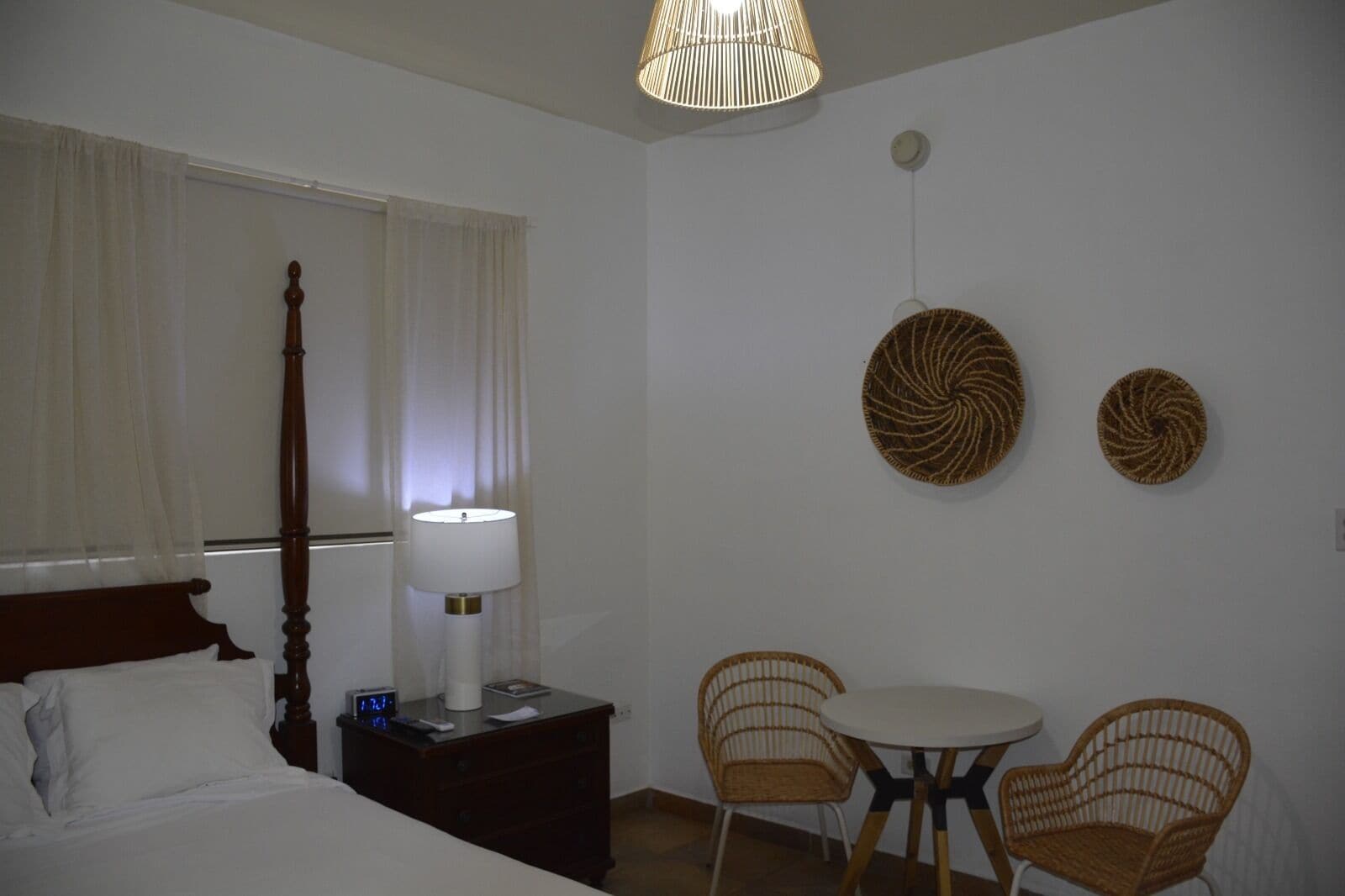 A hotel room with a bed, bedside table, lamp, two wicker chairs, and a round table.