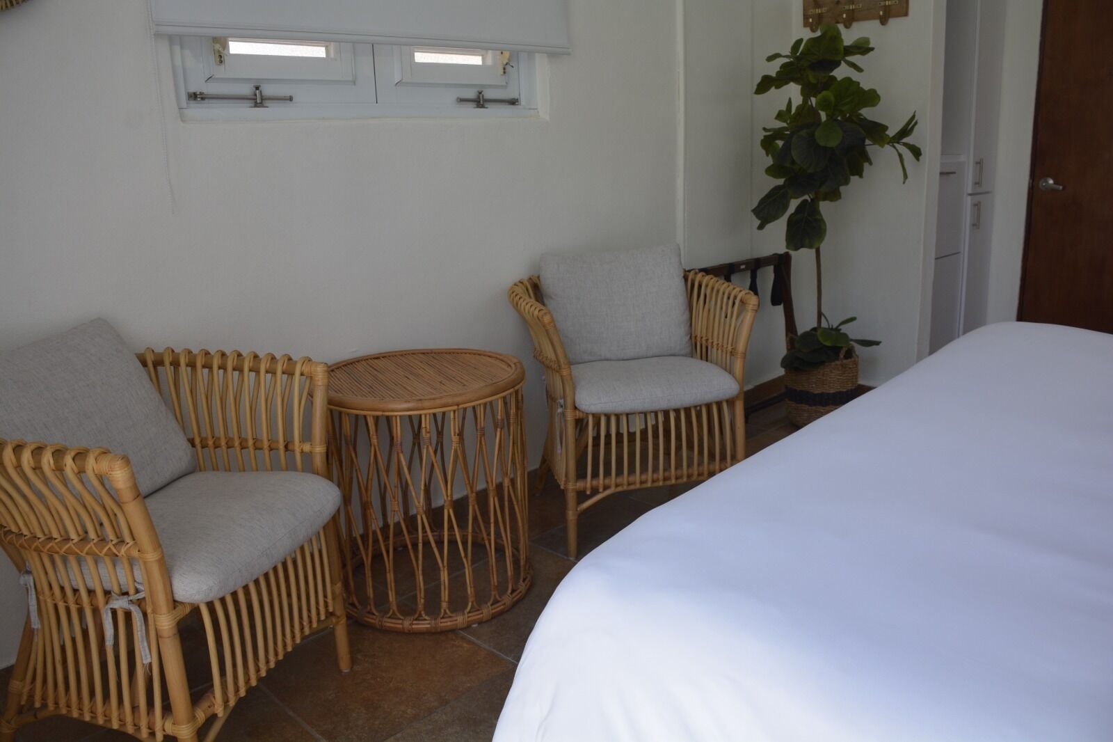 Una habitación con una cama, dos sillones de mimbre, una mesita redonda, una planta en maceta y una ventana con persianas.