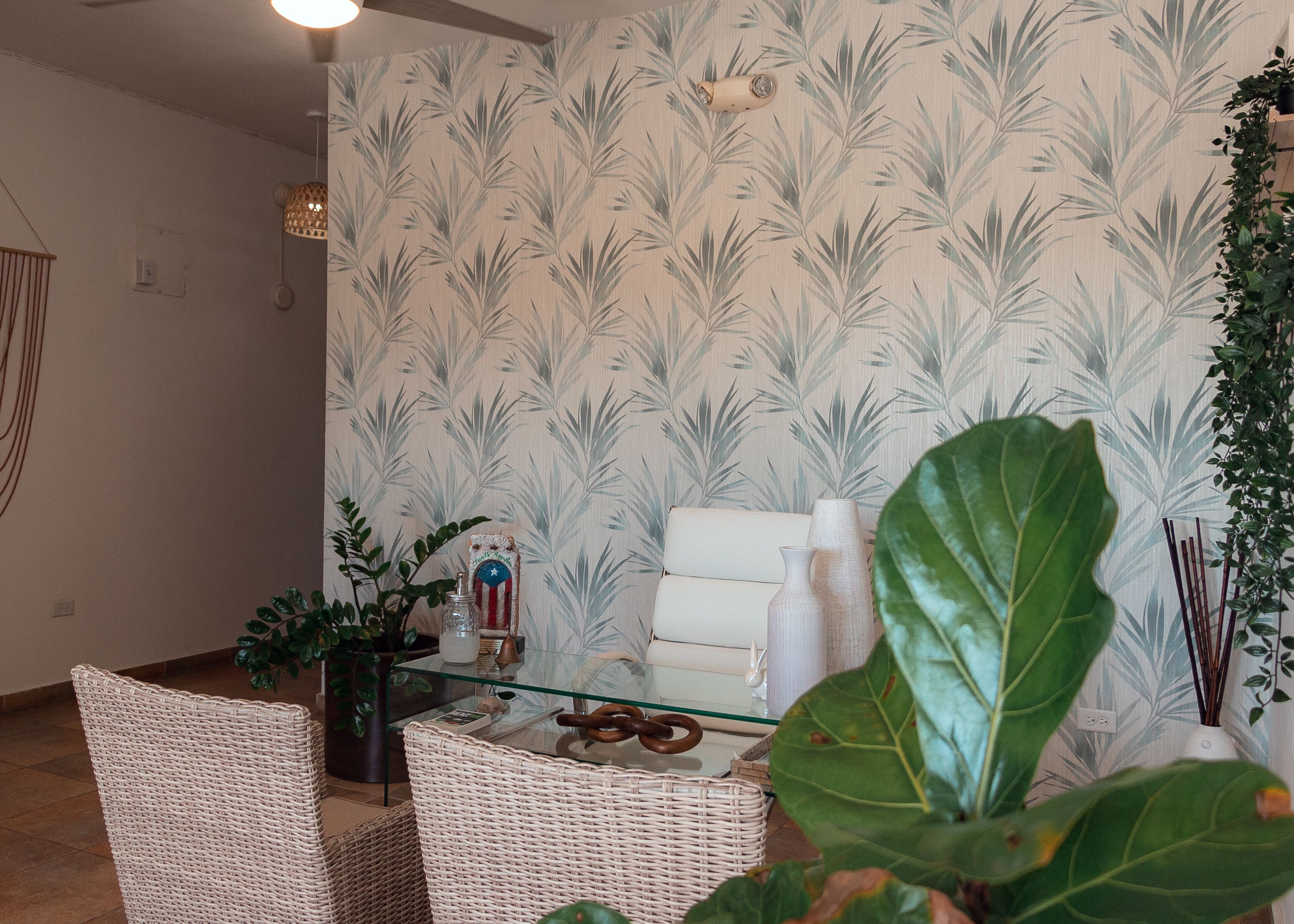 Ein Zimmer mit gemustertem Wandpapier, Korbmöbeln, einem Glastisch und einem Blumentopf.