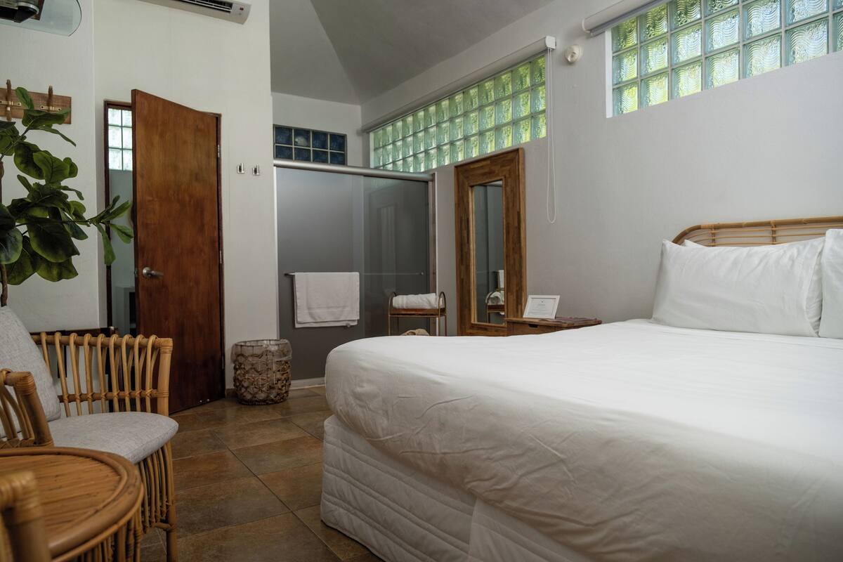 Quarto com cama grande, cadeira de madeira, mesa pequena, espelho e área de chuveiro.