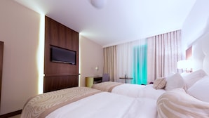 A hotel room with two beds, a TV mounted on the wall, a desk with a chair, and a small table with a lamp. - Hotel Transilvania (Alba Iulia)
