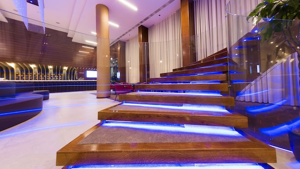 A modern interior with a wooden staircase illuminated by blue lighting, a reception area with seating, and a large plant. - Hotel Transilvania (Alba Iulia)
