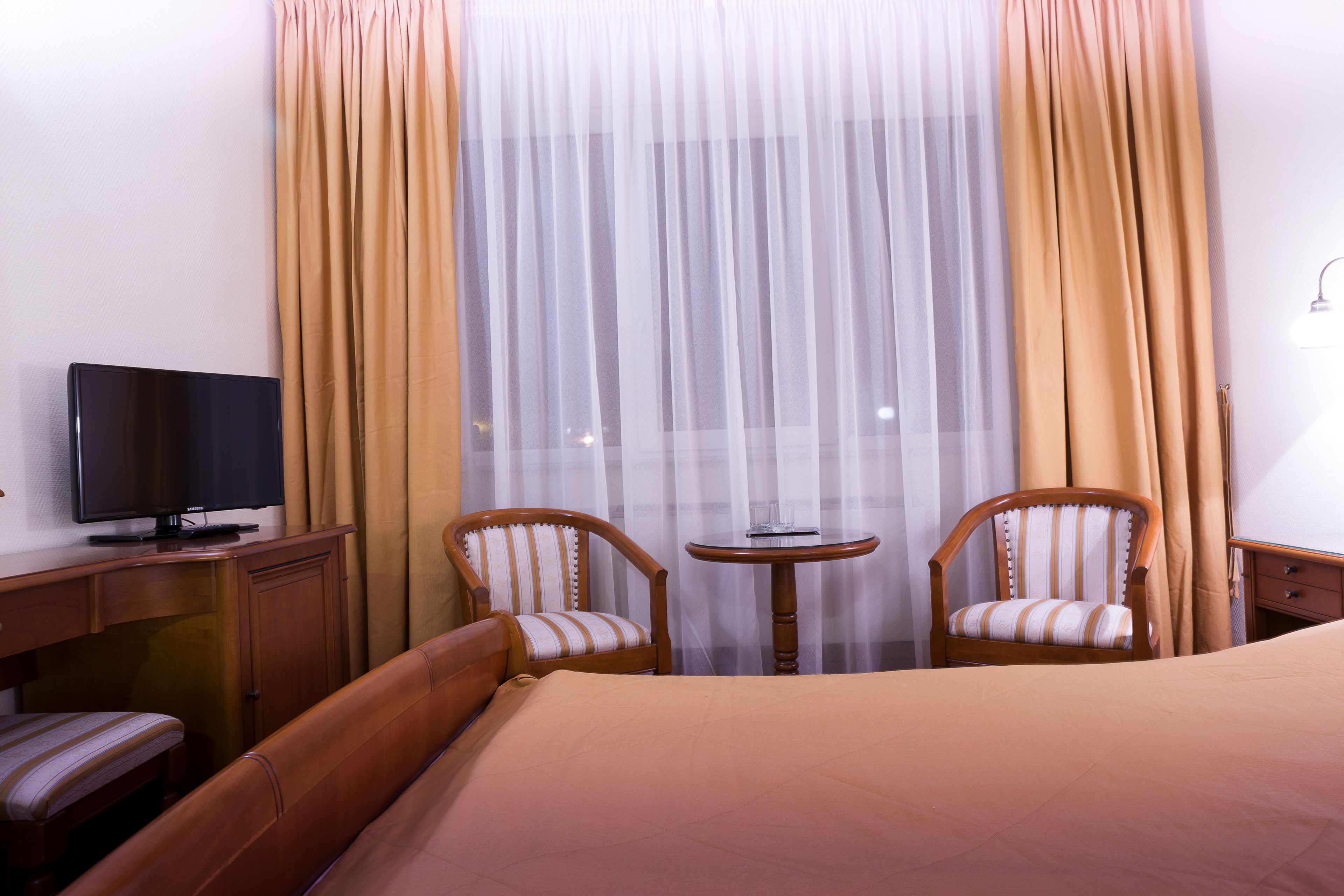Habitación de hotel con cama, dos sillones a rayas, una mesita redonda, televisor sobre un mueble de madera y cortinas traslúcidas.