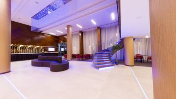A modern lobby with a curved reception desk, a staircase with blue lighting, and a seating area with red chairs.