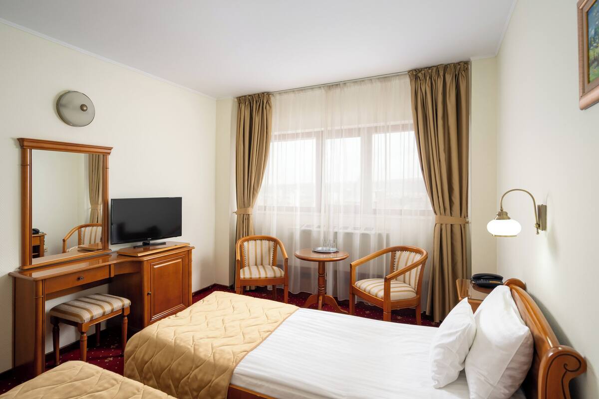 A hotel room with a bed, a desk with a television, two chairs, a small table, and a window with curtains.