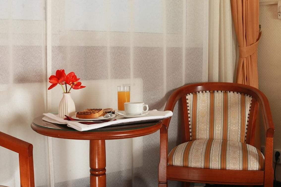 Une petite table en bois, sur laquelle se trouvent un vase blanc, des fleurs rouges, une assiette de pâtisseries, une tasse et un verre de jus d’orange. Une chaise à rayures est placée à côté de la table.