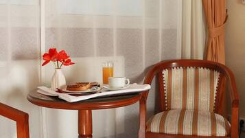 Une petite table en bois, sur laquelle se trouvent un vase blanc, des fleurs rouges, une assiette de pâtisseries, une tasse et un verre de jus d’orange. Une chaise à rayures est placée à côté de la table.