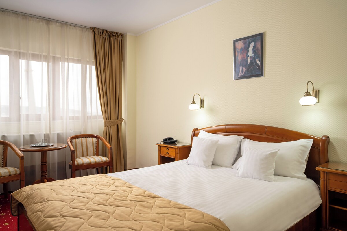 A hotel room with a bed, two bedside tables, a nightstand, a chair, a small table, and a window with curtains.