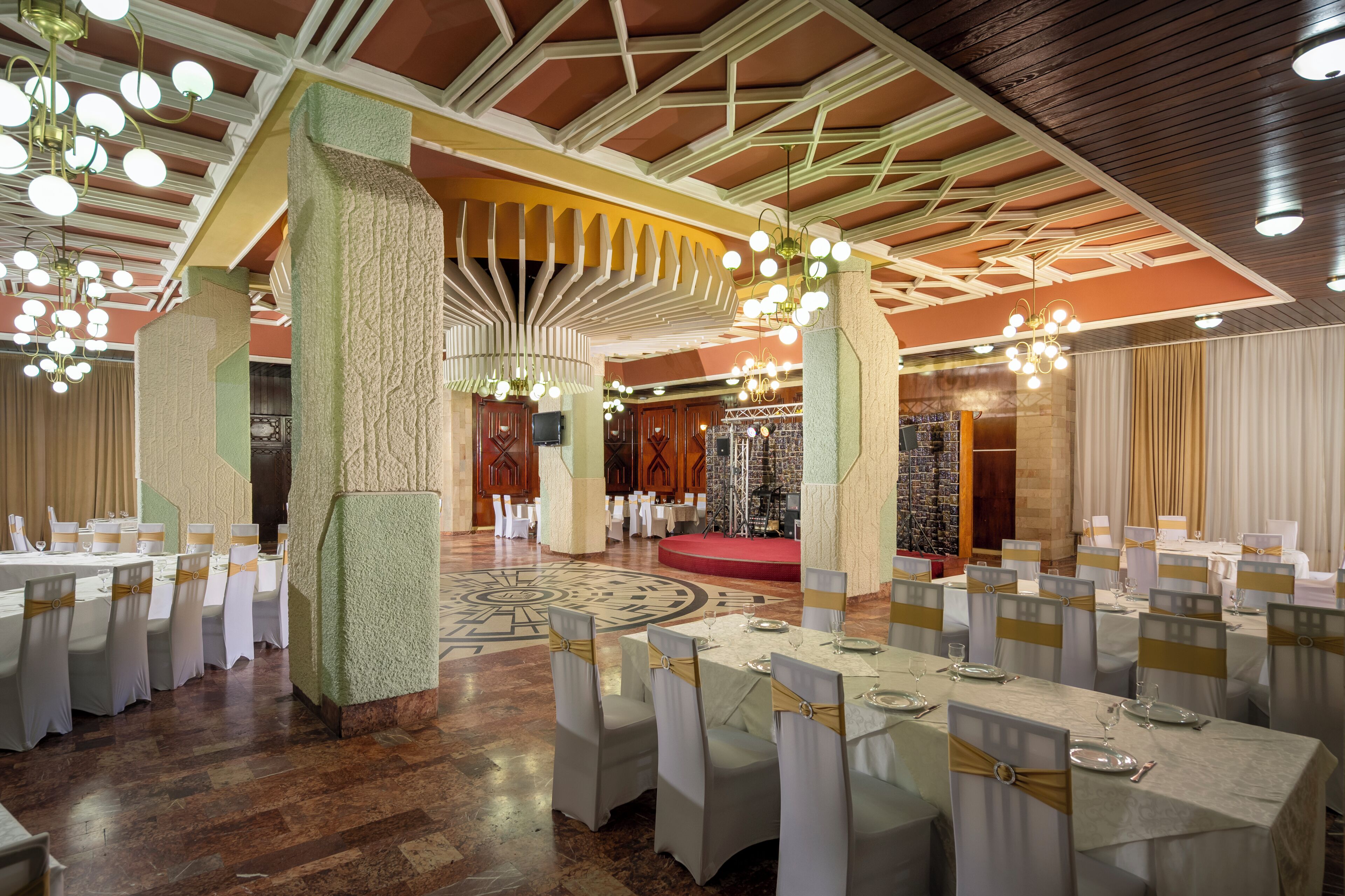 Un salón de banquetes con mesas redondas, sillas blancas y una zona elevada para el escenario.