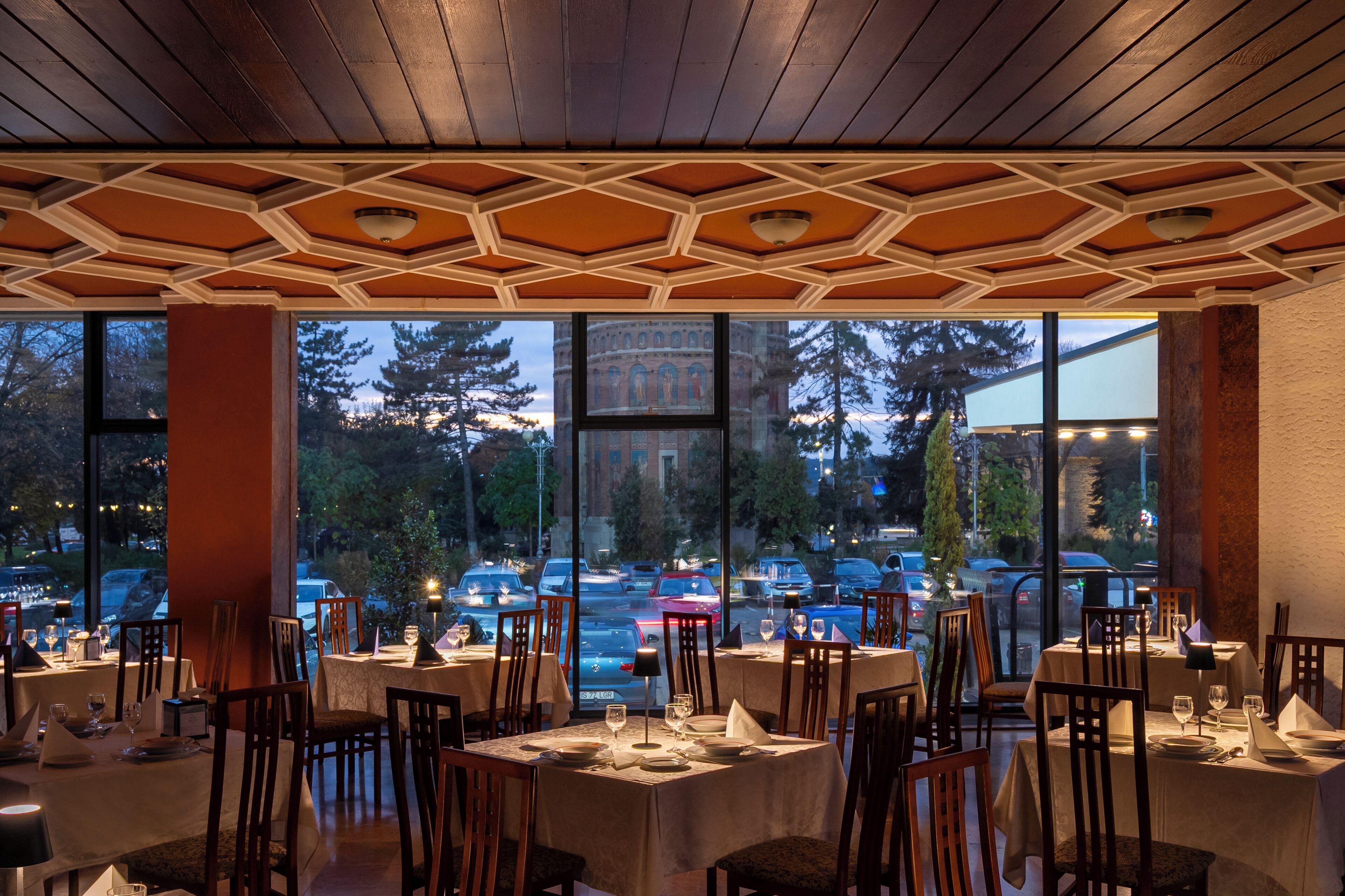 Un ristorante con tavoli apparecchiati, ampie finestre che danno su un parcheggio e alberi.