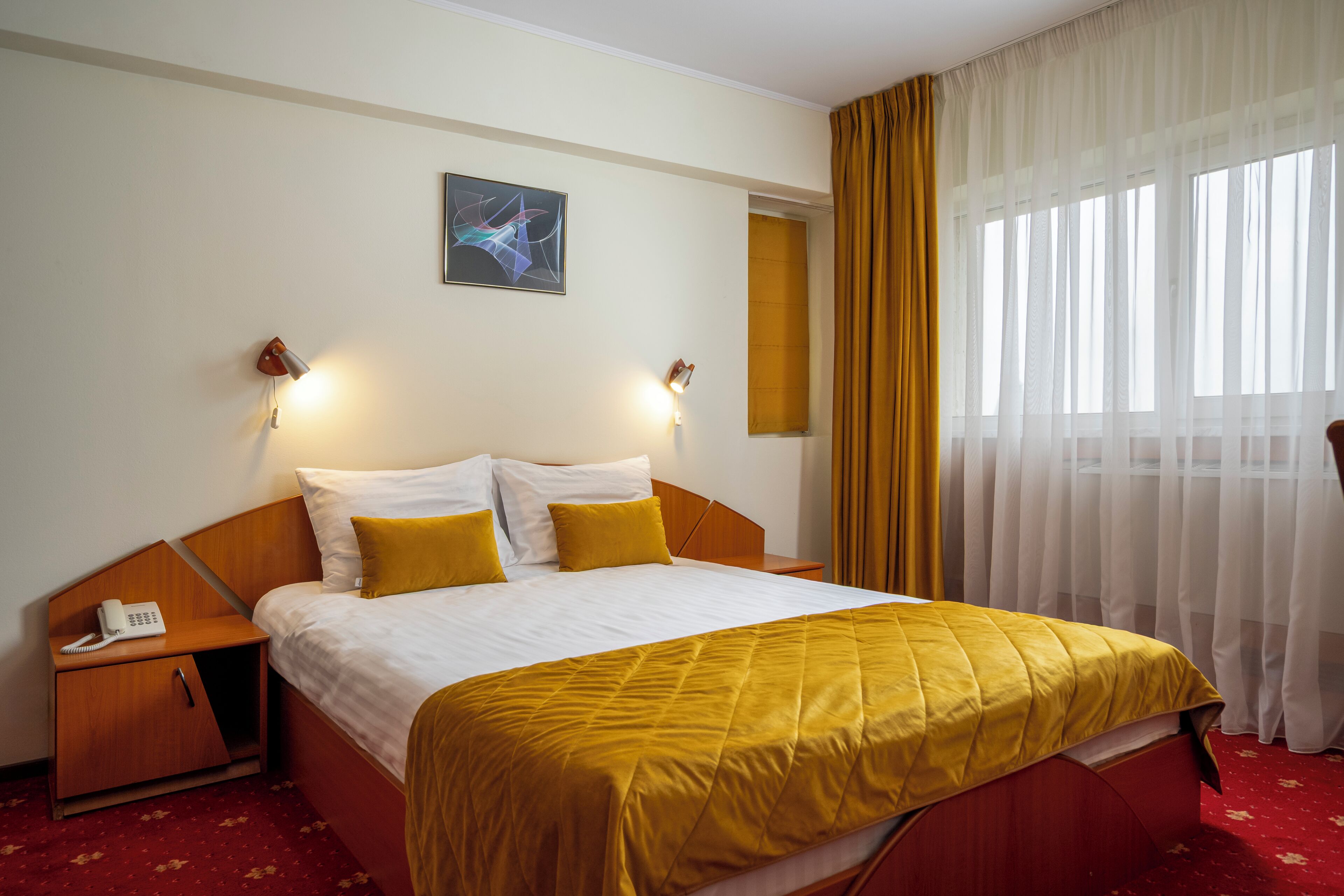 A hotel room with a bed, a bedside telephone, a nightstand, a painting on the wall, and a window with curtains.