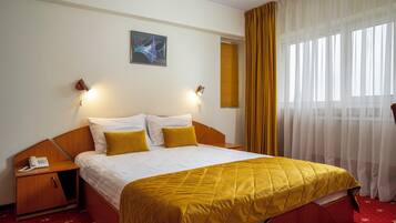 Una camera d'albergo con un letto, un telefono sul comodino, un tavolino, un quadro appeso e una finestra con tende.