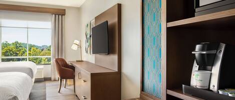 Habitación de hotel con cama, escritorio, silla, televisión, cafetera y vistas al exterior.