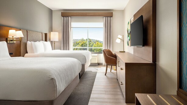 Ein Hotelzimmer mit zwei Betten, einem Schreibtisch, einem Sessel, einem Fernseher und einem großen Fenster mit Vorhängen.