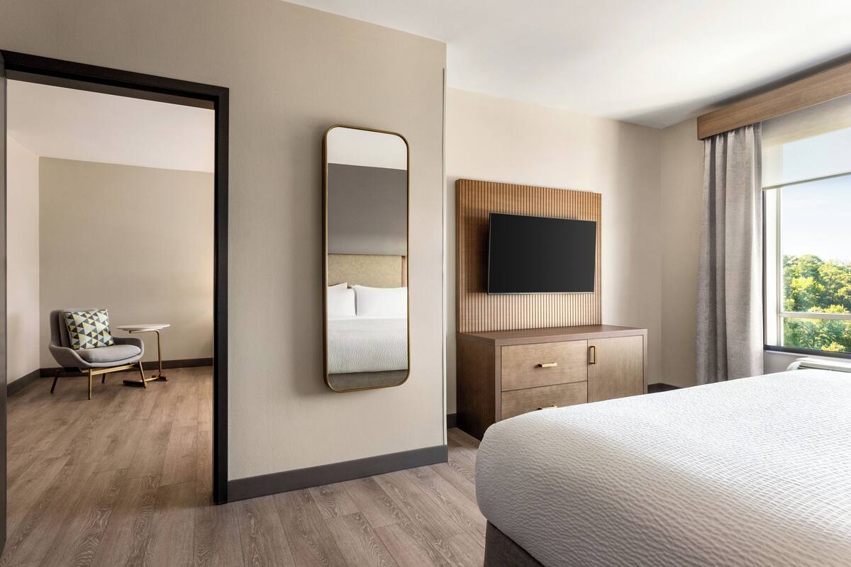 A hotel room with a large bed, a TV mounted on a wooden panel, a small table with a chair, and a mirror on the wall.