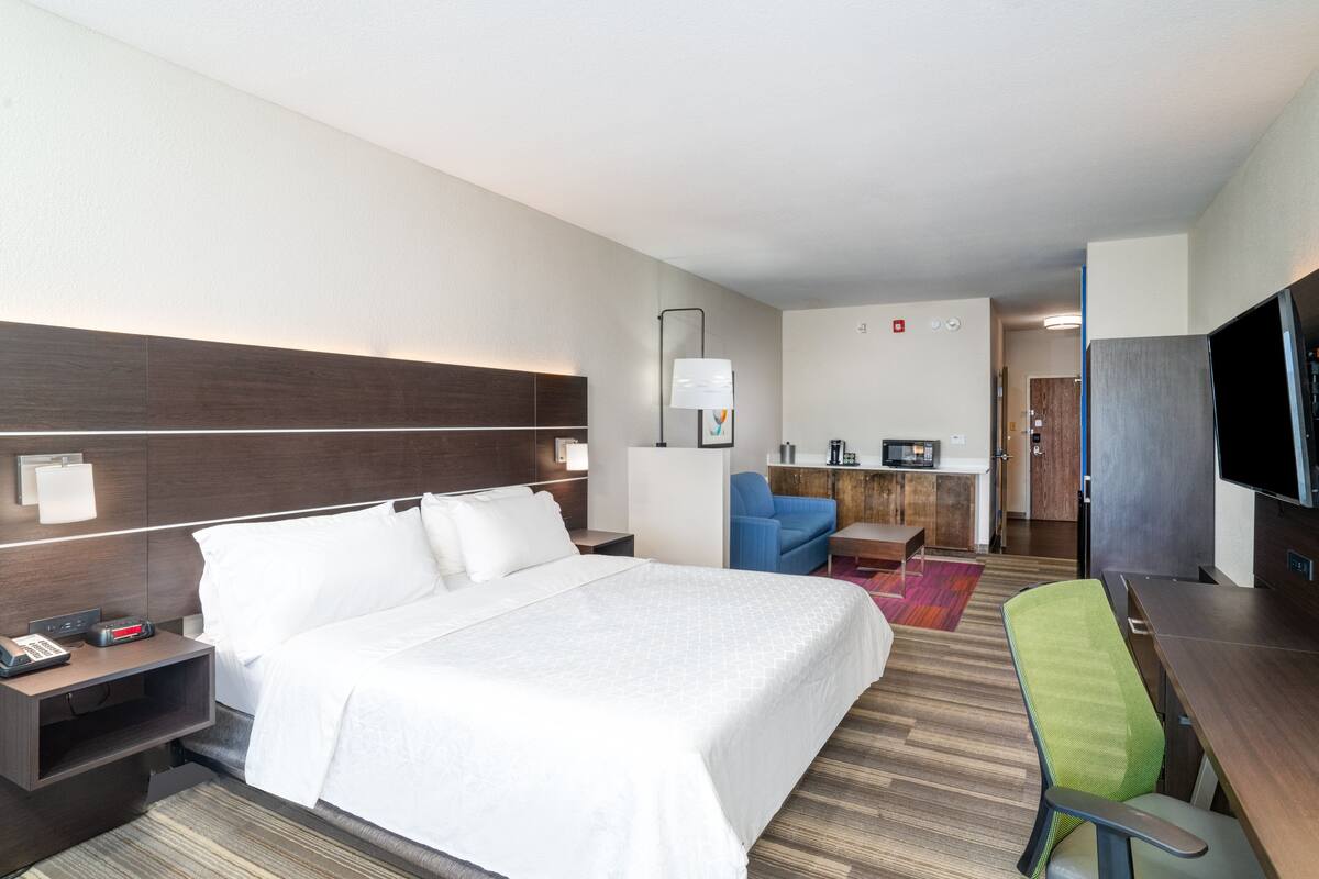 A hotel room with a large bed, a desk, a chair, a television, and a kitchenette.