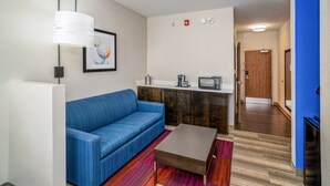A modern living room with a blue sofa, a coffee table, a white lamp, and a kitchenette area with a microwave and toaster. - Holiday Inn Express and Suites Winona North by IHG (Winona)