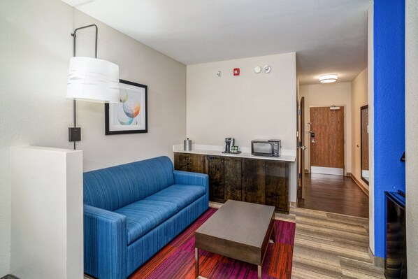 A modern living room with a blue sofa, a coffee table, a white lamp, and a kitchenette area with a microwave and toaster. - Holiday Inn Express and Suites Winona North by IHG (Winona)
