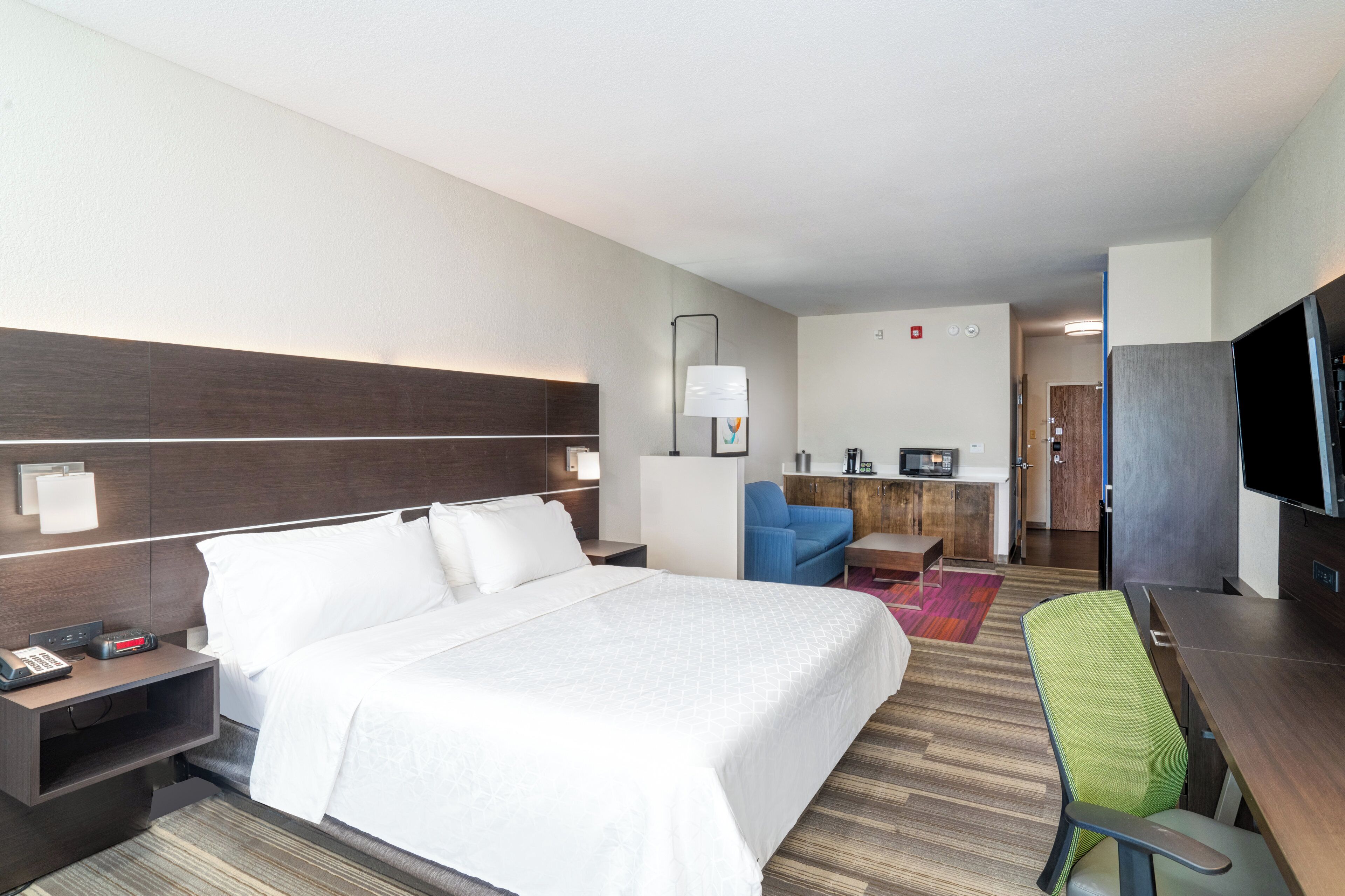 A hotel room with a large bed, a desk, a chair, a television, and a kitchenette.