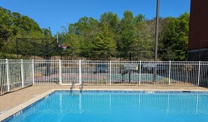 A swimming pool with a white fence, a basketball court, and trees in the background.