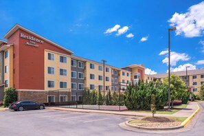 A multi-story hotel with a red and beige facade, a parking lot, and a landscaped area with trees and shrubs.