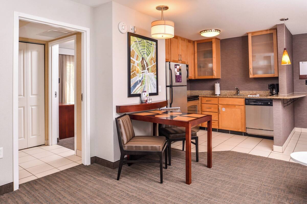 A hotel room with a dining area, a kitchen with wooden cabinets, a refrigerator, and a large window.