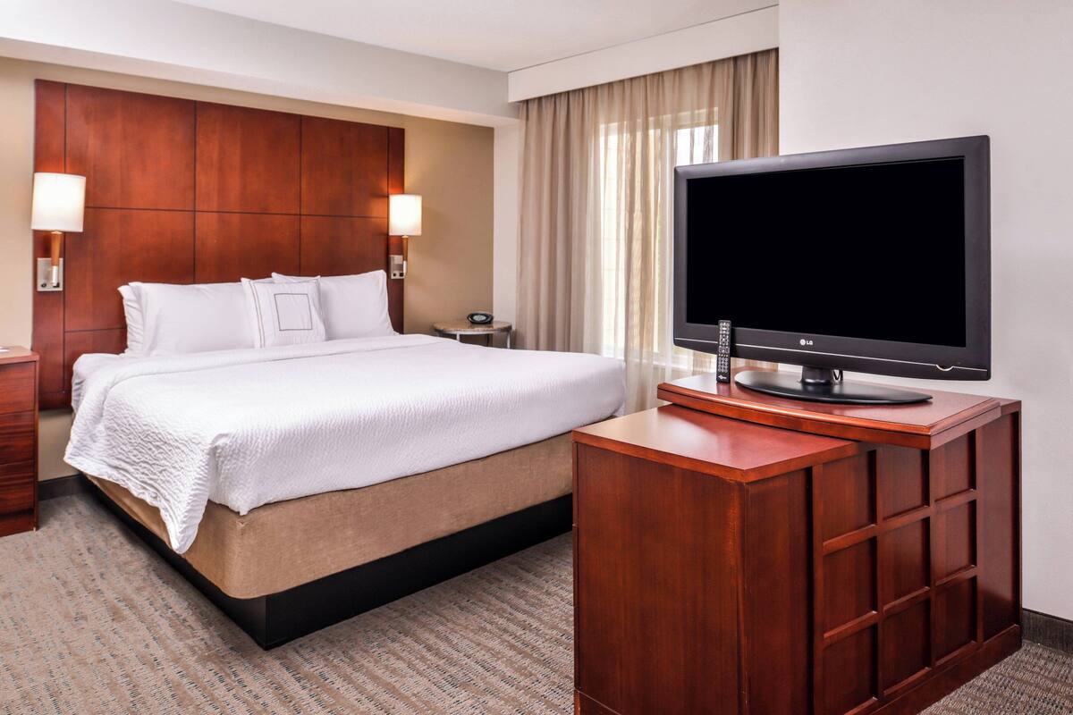 A hotel room with a large bed, a wooden desk with a flat-screen TV, two bedside lamps, and a nightstand.