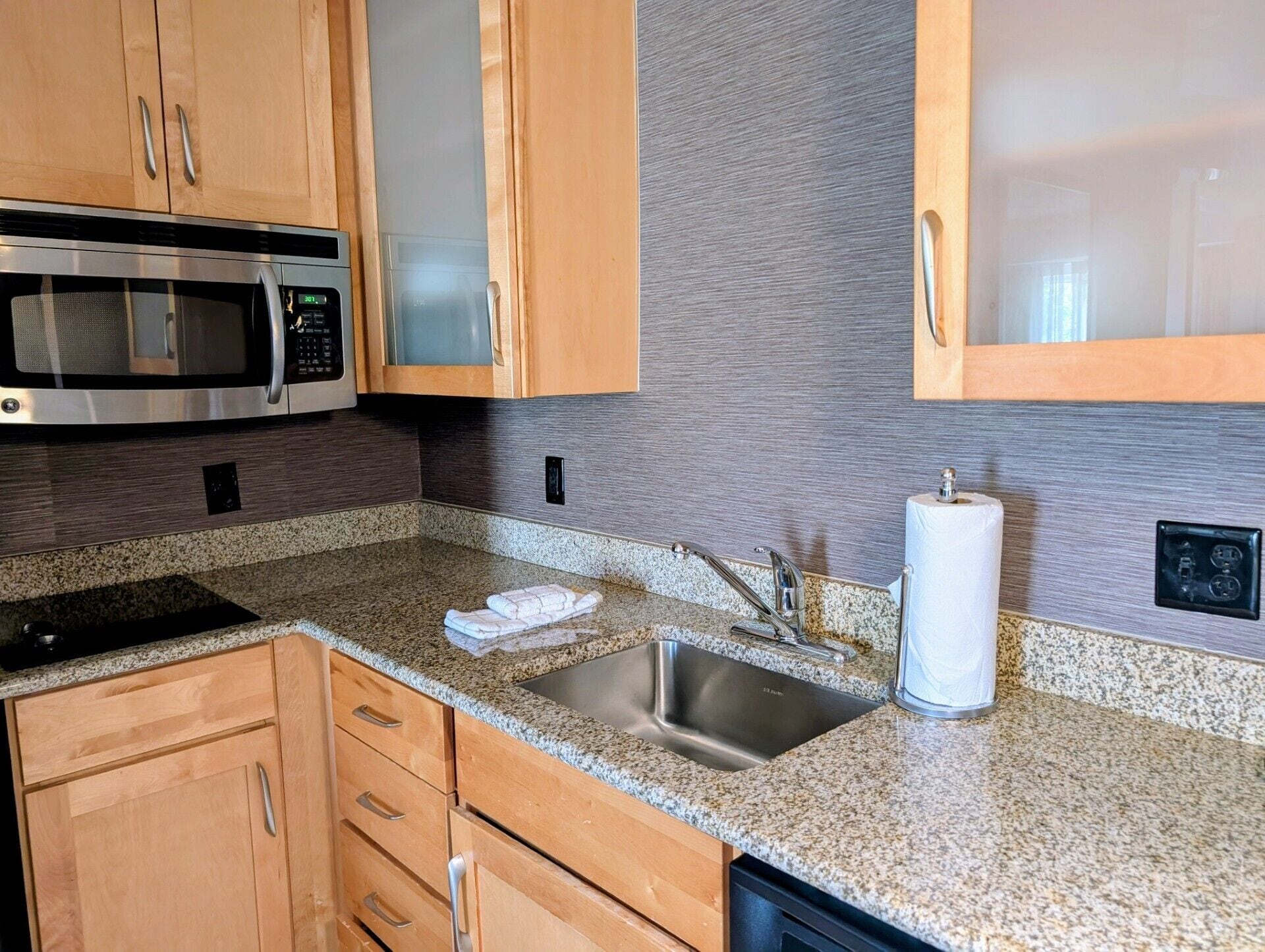 A kitchen with wooden cabinets, a stainless steel sink, granite countertops, a microwave, and a paper towel dispenser.