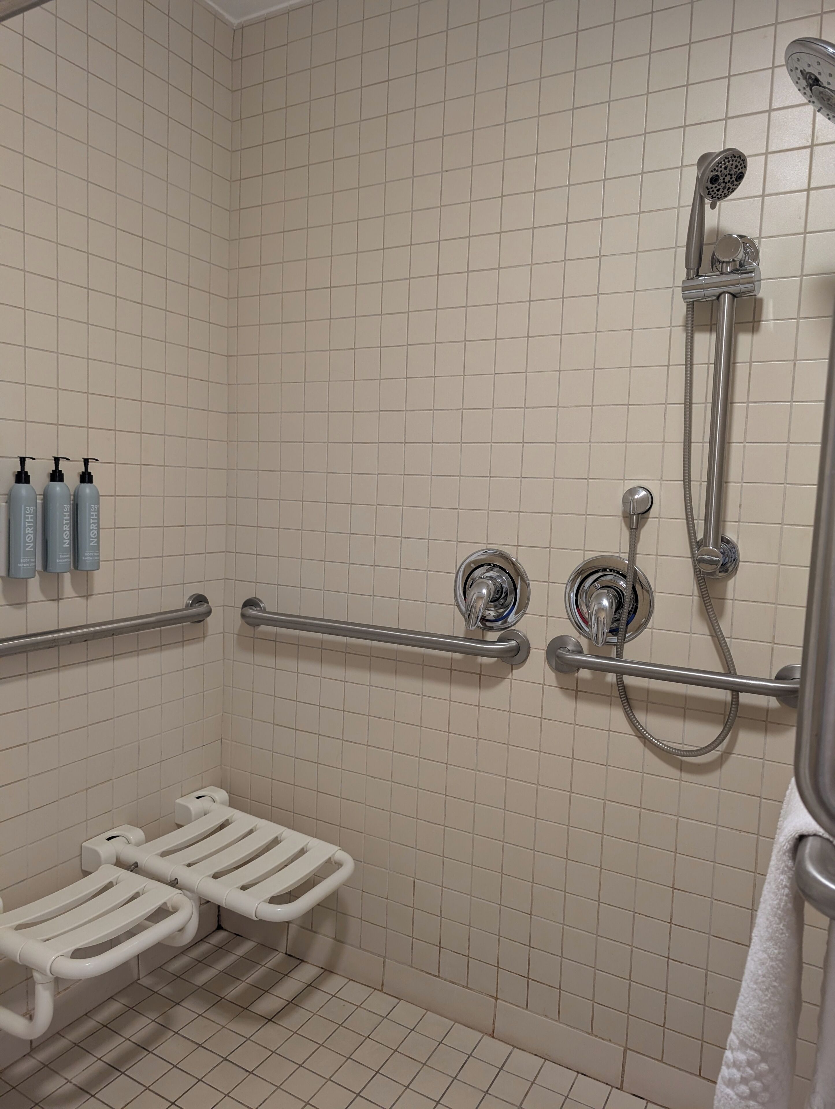 A bathroom with white tiled walls, a shower with a handheld showerhead, grab bars, and a white towel rack.
