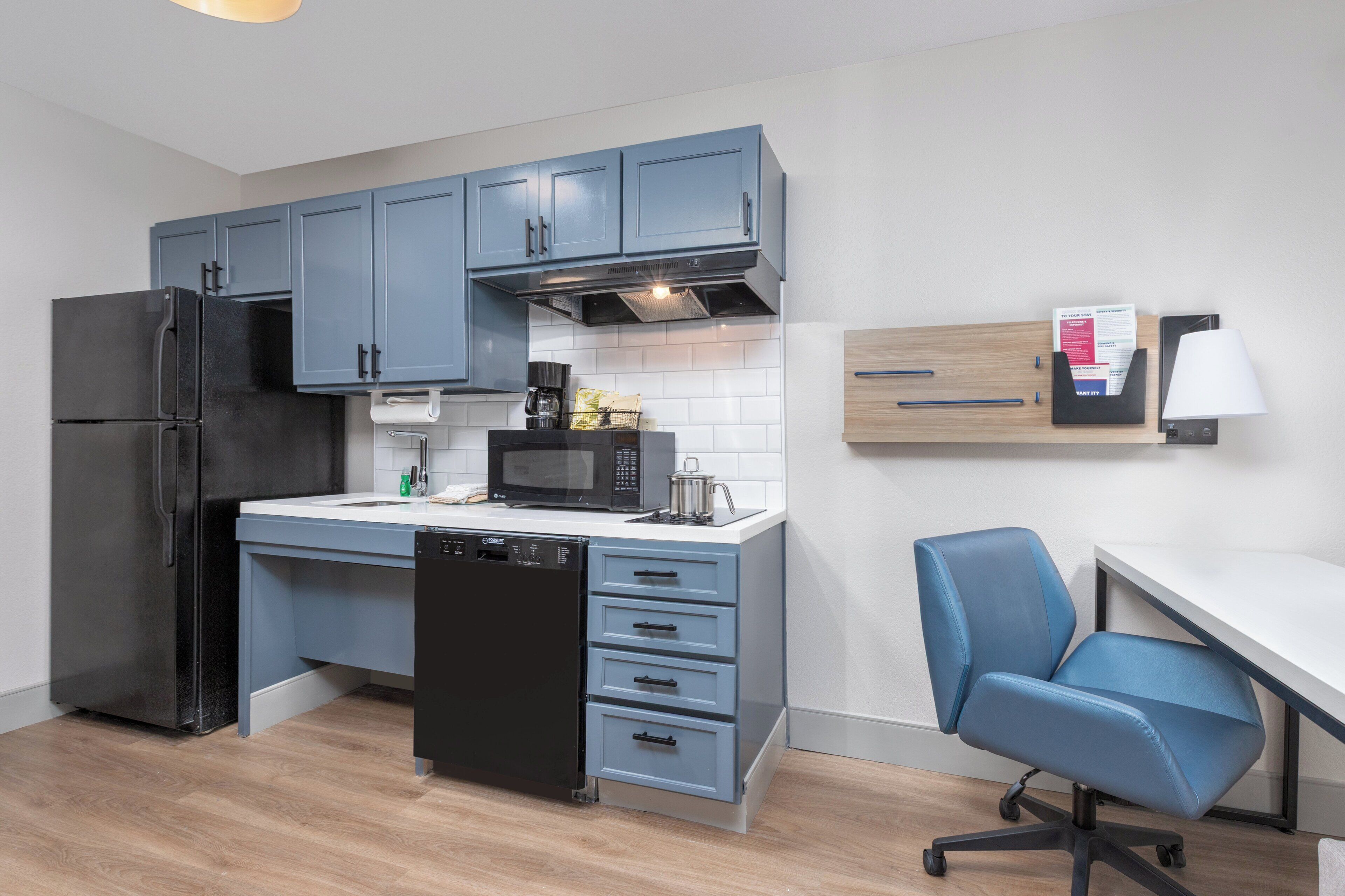 A modern kitchen with blue cabinets, a black refrigerator, a microwave, and a coffee maker. There is a white countertop, a dishwasher, and a blue office chair.
