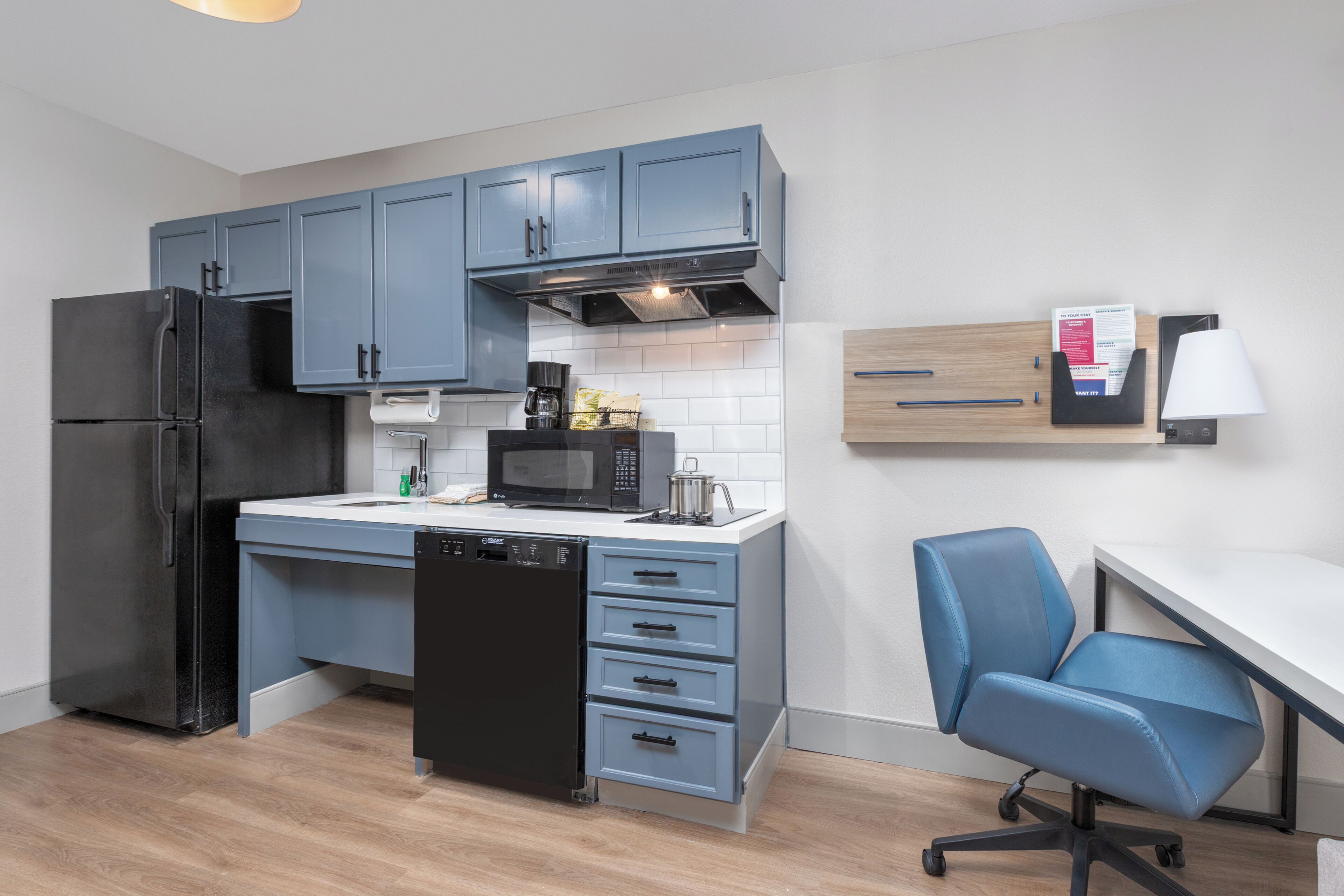 A modern kitchen with grey cabinets, a black refrigerator, a microwave, and a coffee maker. There is a white countertop, a dishwasher, and a blue office chair.