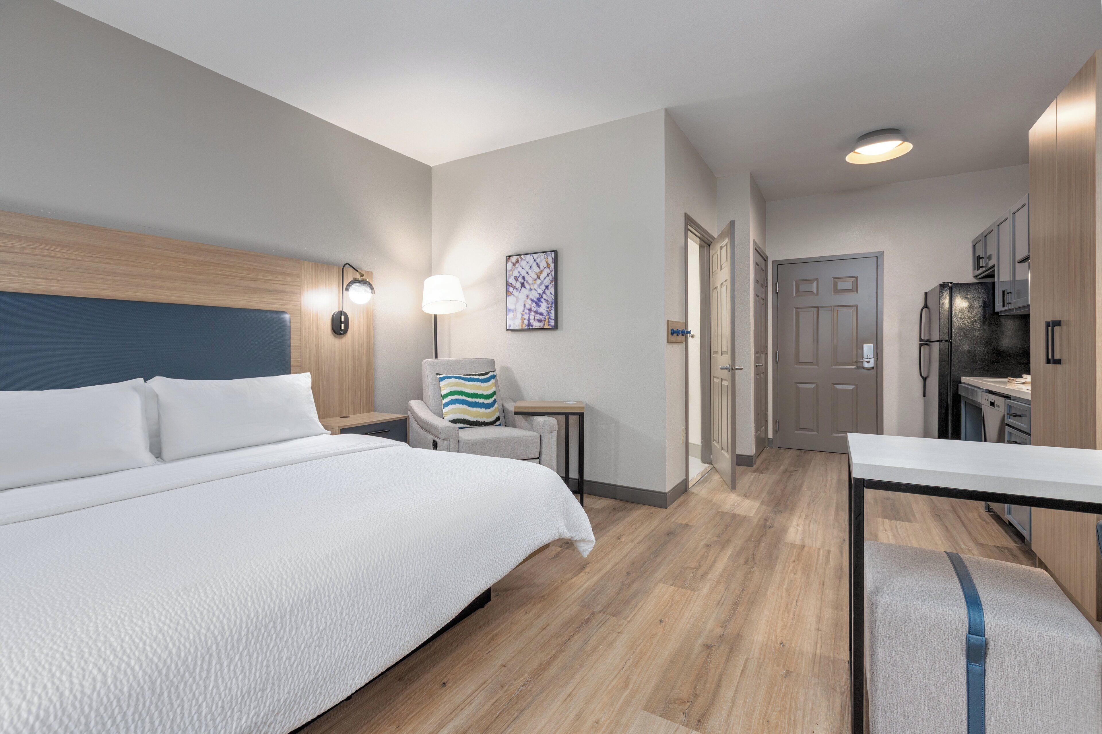 A hotel room with a large bed, a desk, a chair, a lamp, and a refrigerator.