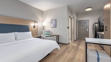 A hotel room with a large bed, a desk, a chair, a lamp, and a refrigerator.