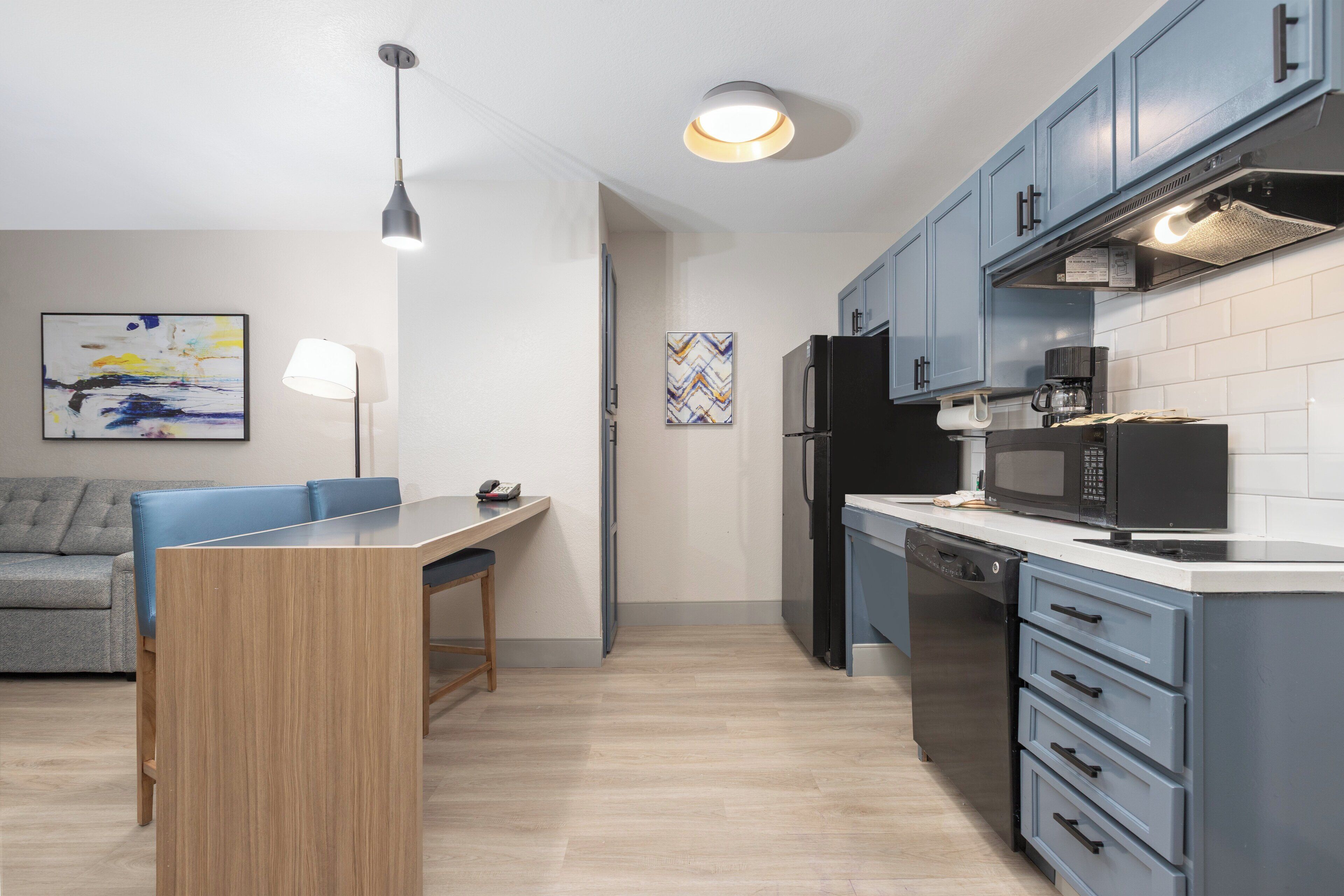 A modern kitchen with light blue cabinets, a black refrigerator, a microwave, and a coffee maker. There is a dining table with chairs and a lamp.