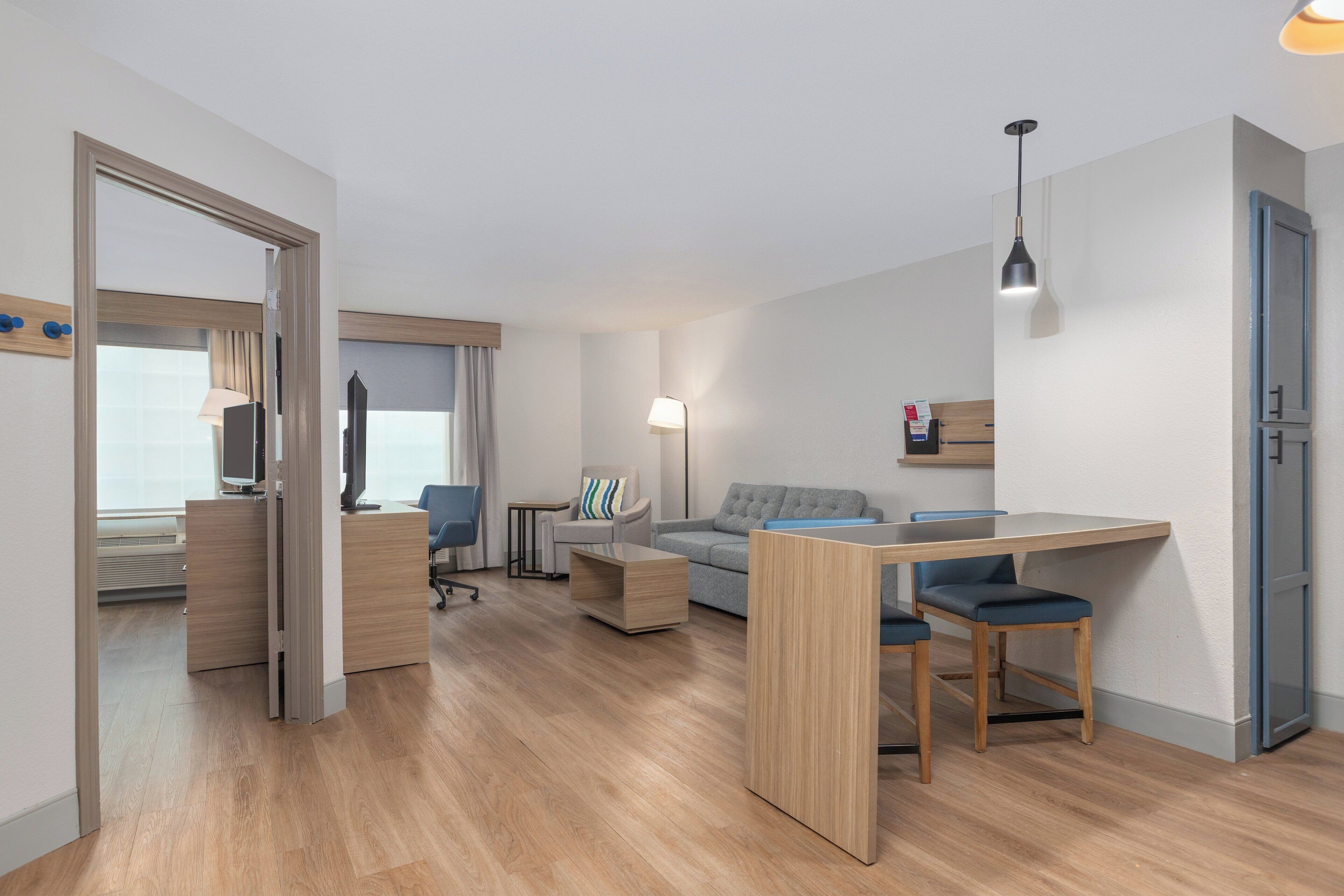 Quarto de hotel moderno com piso de madeira, uma escrivaninha com duas cadeiras, um sofá, uma mesinha e um abajur.