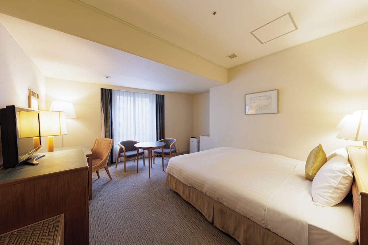 Habitación de hotel con una cama, un escritorio con computadora, una silla, una mesita y un ventanal con cortinas.
