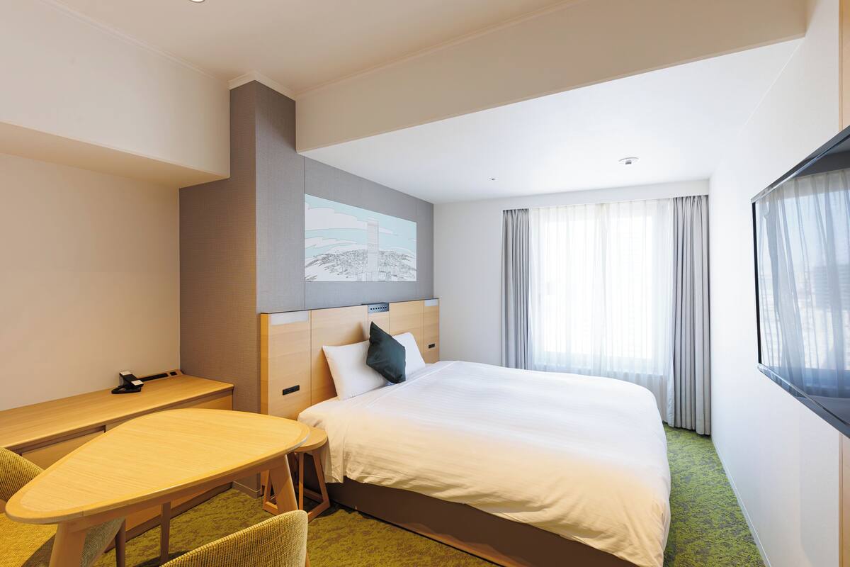Habitación de hotel con cama, escritorio, silla, un cuadro en la pared y una ventana con cortinas.
