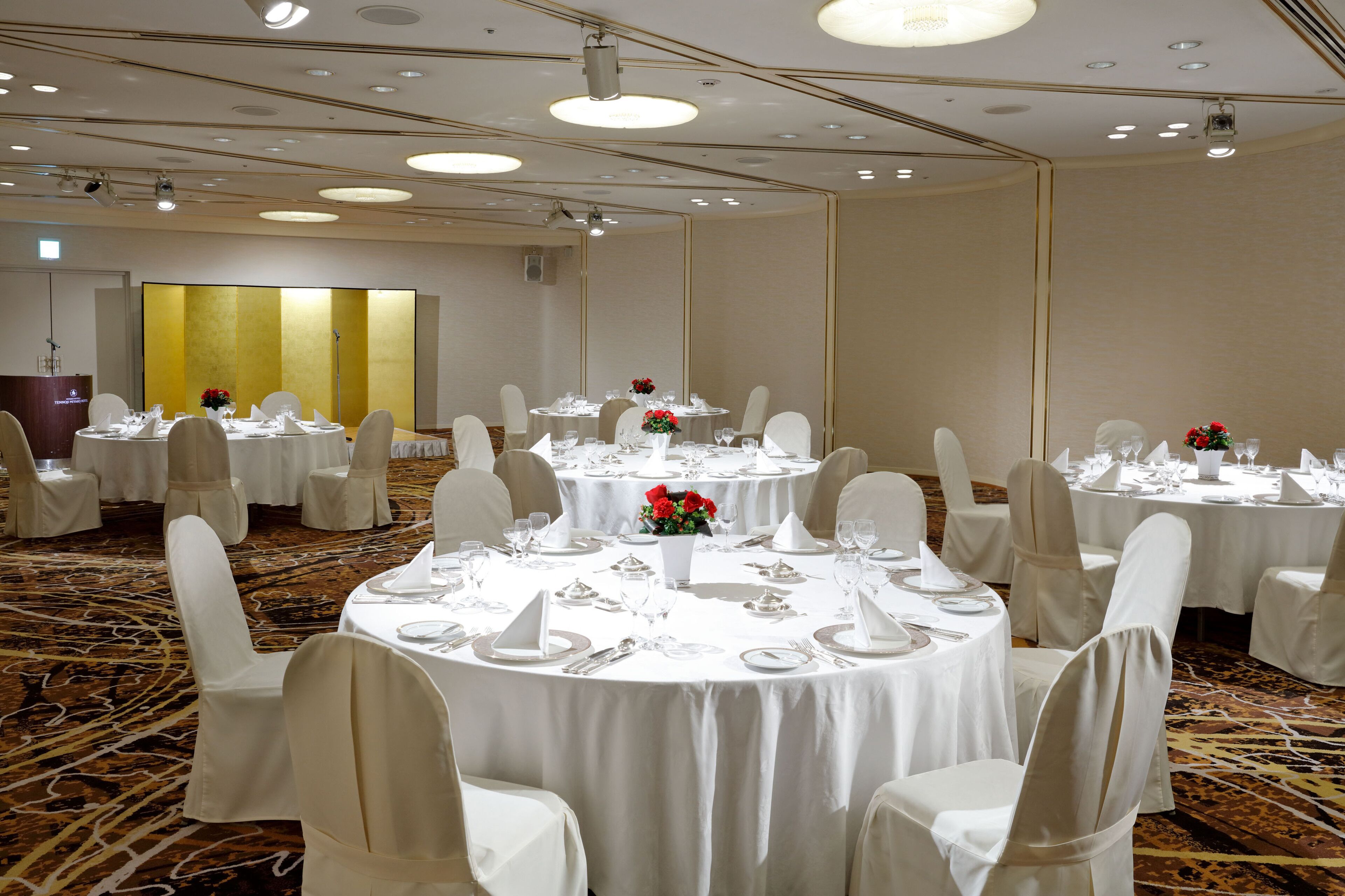 A banquet hall with round tables set for dining, white chairs, and a carpeted floor.