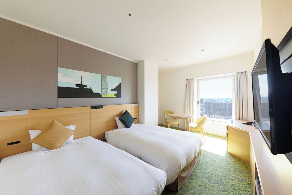 Habitación de hotel con dos camas, un escritorio, televisor y un amplio ventanal con vistas a la ciudad.