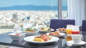 A breakfast table with plates of food, a bowl of salad, and two glasses of juice, overlooking a cityscape.