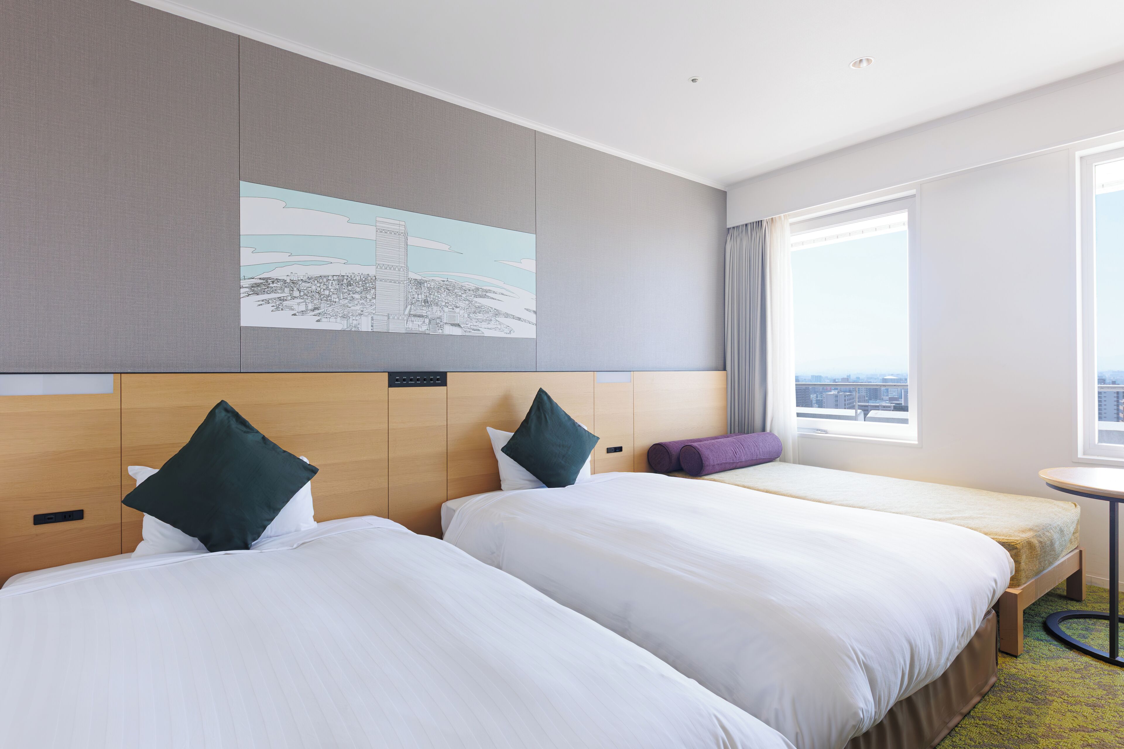 A hotel room with two beds, a small table, a window with a city view, and a painting on the wall.